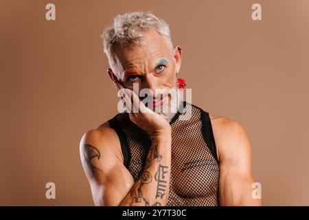 Homme gay mature ennuyé avec des sourcils colorés arc-en-ciel touchant le visage sur fond brun Banque D'Images