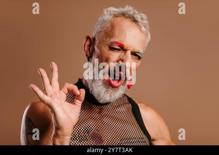 Homme gay mûr ludique avec des sourcils colorés arc-en-ciel faisant des gestes et faisant un clin d'œil sur fond brun Banque D'Images