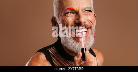 Joyeux homme gay mature avec des sourcils colorés arc-en-ciel appliquant du rouge à lèvres sur fond brun Banque D'Images