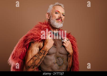 Homme gay mature avec des sourcils colorés arc-en-ciel portant un manteau moelleux sur fond brun Banque D'Images
