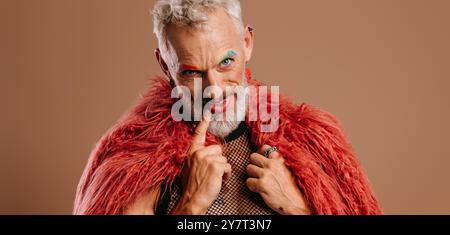 Homme gay mature avec des sourcils colorés arc-en-ciel portant un manteau moelleux sur fond brun Banque D'Images