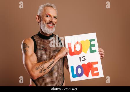 Joyeux homme gay mature avec des sourcils colorés arc-en-ciel tenant la bannière sur fond brun Banque D'Images