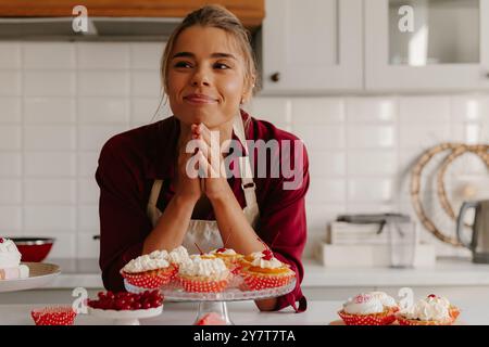 Jeune boulangère attentionnée regardant réfléchi tout en s'appuyant sur la table avec une variété de nourriture sucrée Banque D'Images