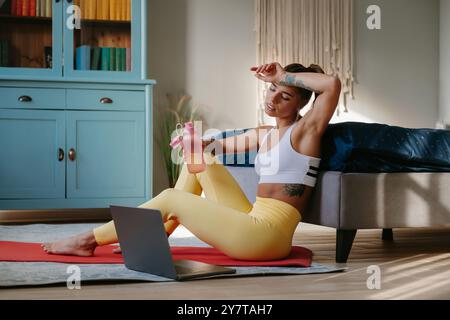 Femme fatiguée dans des vêtements de sport tenant la bouteille d'eau et essuyant la sueur du front tout en se reposant après l'entraînement à la maison Banque D'Images