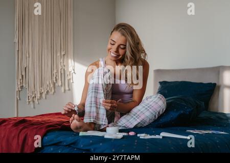 Heureuse jeune femme peignant les ongles des orteils tout en faisant de la pédicure sur le lit à la maison Banque D'Images