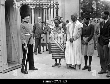 L'uniforme écarlate et la peau d'ours portés par une sentinelle en service à l'extérieur du palais de Buckingham, sont admirés par le roi habillé Akenzua II, Oba du Bénin, sa femme, la reine Ohan et leur fille de 19 ans, la princesse Egbebakhame (à droite), Londres, Angleterre. - L'Oba, dirigeant de 1 000 000 Nigérians, est arrivé en Angleterre hier en tant qu'invité du British Council. Avec lui, il apporta un message de bonne volonté au roi. - 12 août 1950 Banque D'Images