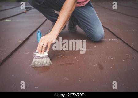 Gros plan d'un homme accroupi en peignant un toit. Sa main tient un pinceau, appliquant soigneusement une couche de peinture fraîche sur la surface. L'accent est mis sur th Banque D'Images