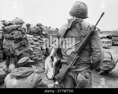 Homme pour toutes les saisonsA marine américaine avec un fusil M-16 sur une épaule et sa guitare sur l'autre, alors qu'il attend à la piste d'atterrissage à Khe Sanh pour un vol hors de la forteresse assiégée. Vietnam du Sud - 21 février 1968 Banque D'Images