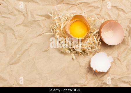 Oeuf cassé sur paille. Coquille d'œuf et plumes d'oiseau sur la table. Fond brun de papier froissé. Pose à plat Banque D'Images
