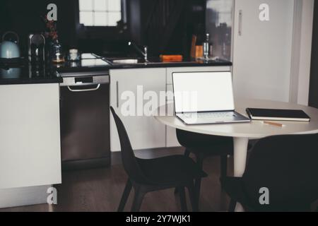 Un espace de travail de cuisine minimaliste avec un ordinateur portable élégant sur une table ronde, accompagné d'un cahier et d'un crayon. Décor moderne noir et blanc, lignes épurées, Banque D'Images