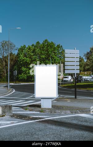Plan vertical de la maquette d'un panneau d'affichage vierge et de panneaux de direction vides à un rond-point de la ville. Ciel dégagé, verdure et rue visible. Parfait pour Banque D'Images