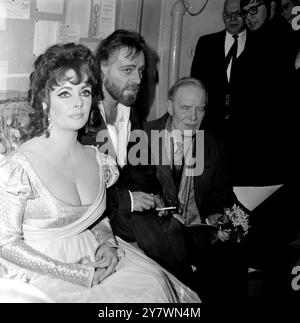 N'ayant pas dit un mot tout au long de la performance ... actrice Elizabeth Taylor est assis silencieusement dans le vestiaire comme mari Richard Burton parle à la presse ... après l'ouverture de la nuit du Docteur Faustus à l'Oxford Playhouse . Les deux stars ont donné leurs services gratuitement pour la semaine de cette pièce , dont les profits sont en aide à la Oxford University Dramatic Society . 15 février 1966 Banque D'Images