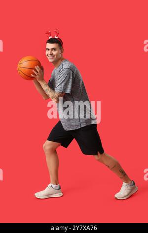 Jeune homme heureux dans des cornes de renne jouant au basket-ball sur fond rouge Banque D'Images