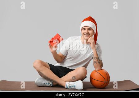 Jeune homme dans le chapeau de Père Noël tenant la boîte cadeau près du basket-ball sur fond gris Banque D'Images