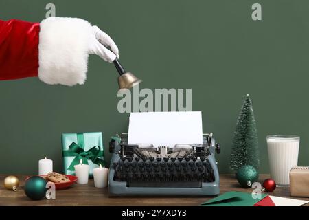 Main du Père Noël avec cloche de Noël, biscuits, verre de lait et machine à écrire à la table en bois Banque D'Images