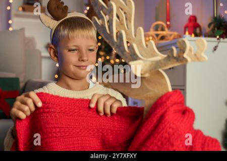 Petit garçon enveloppant renne en bois avec écharpe à la maison la veille de Noël, gros plan Banque D'Images