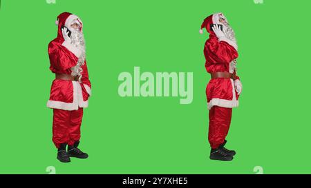 Santa parle sur appel téléphonique en studio, en utilisant la ligne de smartphone pour avoir le chat à distance pendant les vacances de noël. Saint Nick cosplay en costume discutant avec les gens sur fond d'écran vert du corps entier. Banque D'Images