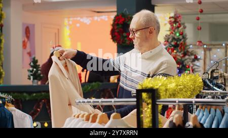 Homme âgé fatigué parcourant les vêtements dans le magasin de mode festif, à la recherche de cadeaux de famille, fatigué des achats de Noël. Client senior choisissant blazer élégant pour neveu Banque D'Images