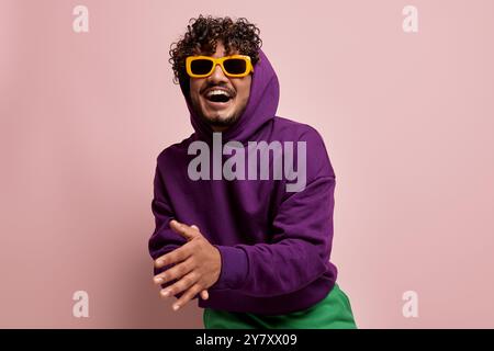 Heureux jeune homme indien dans des lunettes de soleil élégantes souriant et faisant des gestes sur fond rose Banque D'Images