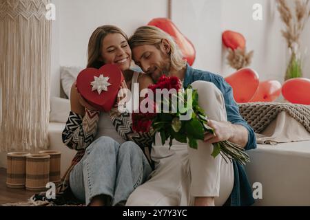 Couple d'amour embrassant et tenant des roses et une boîte cadeau tout en célébrant la Saint-Valentin à la maison confortable Banque D'Images