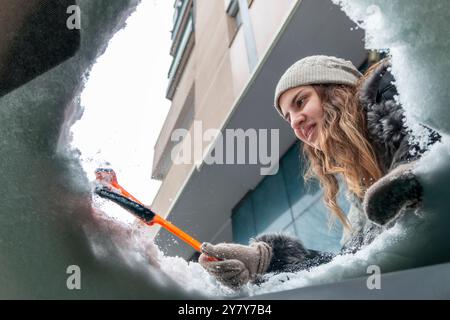 Jeune femme utilisant une brosse à neige pour enlever la neige du pare-brise de sa voiture. Concept de météo hivernale, entretien de voiture, et être préparé pour con neigeux Banque D'Images