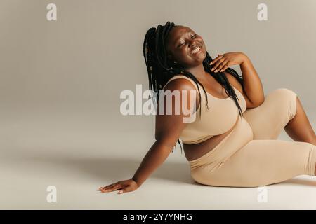 Joyeuse plus courbe femme africaine dans des vêtements de sport rayonnant l'amour de soi tout en étant assis sur fond de studio Banque D'Images