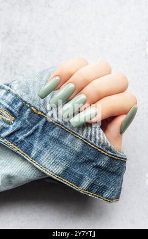 Une main présentant de longs ongles verts élégants est placée sur une manche en denim. L'arrière-plan est une texture douce et neutre qui améliore la couleur des ongles. Banque D'Images