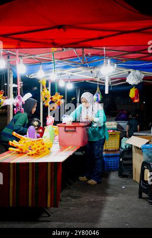 Une femme vend des jouets au marché. Le marché est illuminé de lumières. La femme porte une chemise verte Banque D'Images