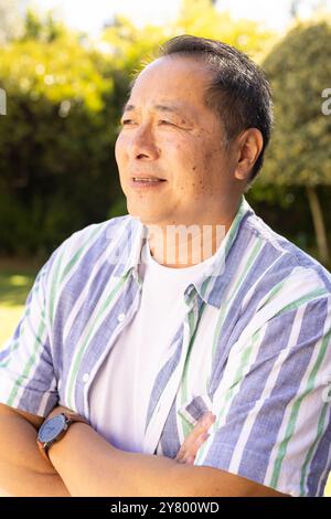 Homme d'âge moyen asiatique souriant debout à l'extérieur, croisant les bras et regardant loin, profitant d'une journée ensoleillée Banque D'Images