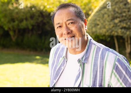Souriant homme d'âge moyen asiatique en chemise rayée profitant d'une journée ensoleillée à l'extérieur dans le jardin Banque D'Images
