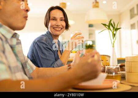 À la maison, souriante femme d'âge moyen appréciant le repas avec la famille, tenant le verre à la table à manger Banque D'Images