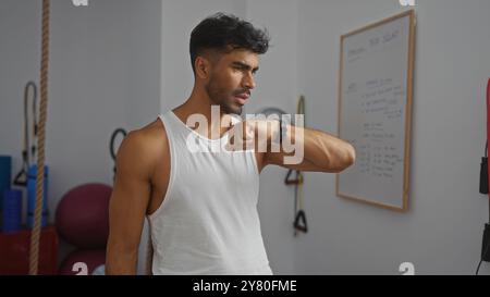 Bel homme dans une salle de sport portant un débardeur blanc vérifiant sa smartwatch tout en étant entouré d'équipements de fitness et d'accessoires de gymnastique. Banque D'Images