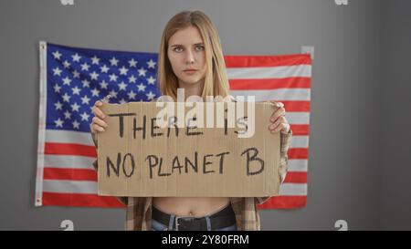 Jeune femme préoccupée tenant une affiche de protestation environnementale au bureau avec le drapeau américain en toile de fond. Banque D'Images