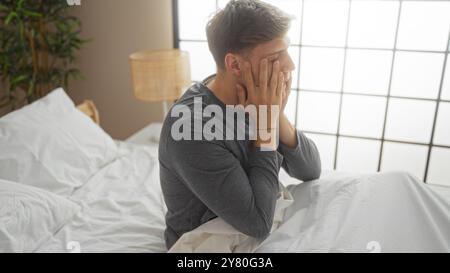 Jeune homme assis sur un lit dans une chambre moderne semblant frustré sur un fond de mur blanc Banque D'Images