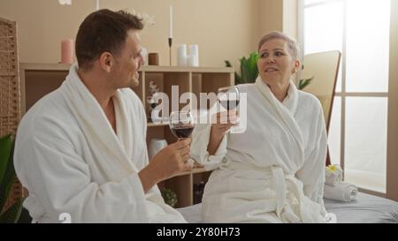 Homme et femme portant des peignoirs blancs dégustant du vin ensemble dans une salle de spa intérieure à l'atmosphère chaleureuse et intime Banque D'Images