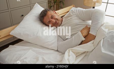 Homme allongé dans le lit avec une expression douloureuse dans une chambre lumineuse avec des draps blancs, mettant en vedette un adulte caucasien d'âge moyen dans un cadre de maison intérieur. Banque D'Images