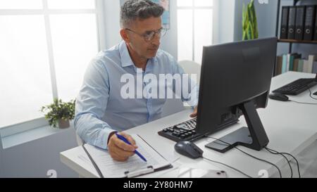 L'homme d'âge moyen travaille à un ordinateur dans un bureau bien éclairé, écrivant sur des documents avec concentration. Banque D'Images
