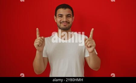 Jeune homme hispanique posant sur un fond rouge isolé pointant vers le haut avec les deux index, portant un t-shirt blanc, souriant avec confiance Banque D'Images