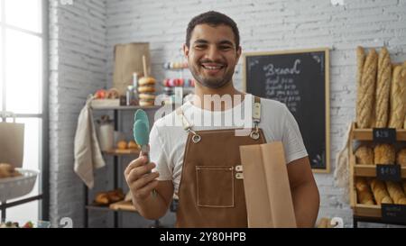 Jeune homme tenant des pinces et un sac en papier souriant dans une boulangerie remplie de pains et pâtisseries assortis Banque D'Images