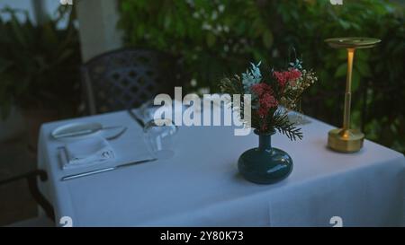 Salle à manger en plein air à majorque, en espagne, avec une nappe blanche, un vase avec des fleurs, des tables élégantes et une lampe dorée au milieu d'une végétation luxuriante Banque D'Images