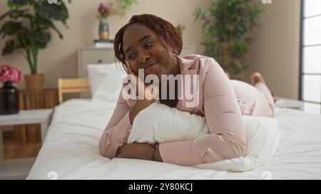 Belle jeune femme afro-américaine se relaxant sur le lit dans une chambre confortable, souriant paisiblement tout en étant allongée sur des oreillers blancs dans un cadre accueillant et lumineux Banque D'Images