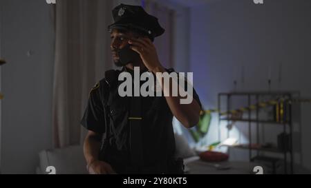 Un homme afro-américain concentré en uniforme de police communique sur un téléphone portable dans une scène de crime intérieure faiblement éclairée. Banque D'Images