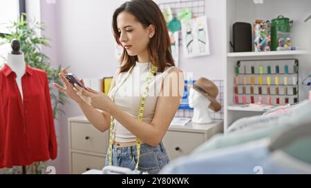 Une jeune femme tailleur vérifie le téléphone dans un atelier de couture coloré avec mannequin et tissu. Banque D'Images