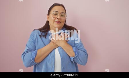 Une femme hispanique d'âge moyen avec des lunettes, exprimant de la gratitude ou des émotions sincères, isolée sur un fond rose. Banque D'Images