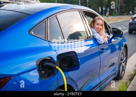 Femme blonde se détend dans son véhicule électrique pendant qu'il se recharge à une borne de recharge de la ville. Banque D'Images