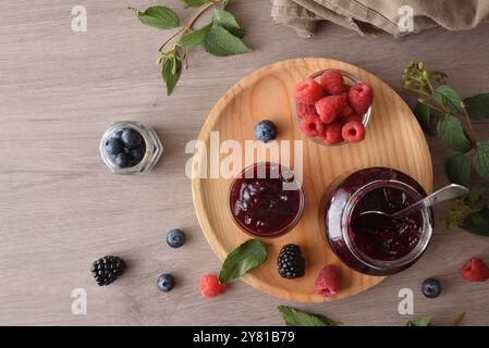 Pots de confiture de fruits de forêt sur une assiette en bois sur une table avec des fruits et des feuilles. Vue de dessus. Banque D'Images