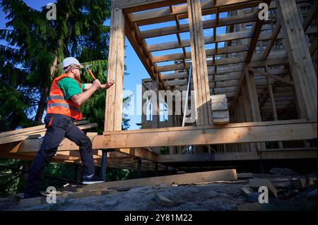 Charpentier construisant une maison à ossature en bois de deux étages près de la forêt. Homme barbu martelant des clous tout en portant un casque de protection et un gilet de construction. Concept de construction écologique moderne. Banque D'Images