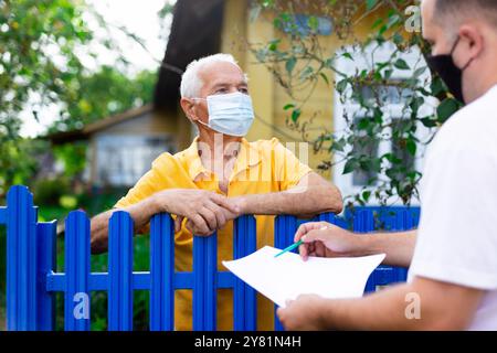 Homme senior en masque de protection communiquant avec le représentant de la compagnie d'assurance tout en se tenant à la clôture de sa maison de campagne Banque D'Images