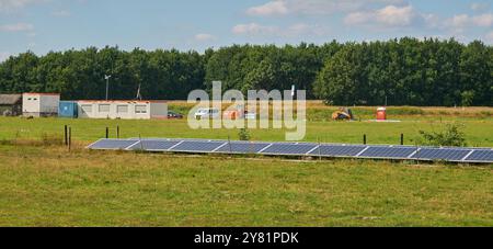 chantier pour une ferme d'énergie solaire en plein soleil à côté d'une autoroute, avec des arbres et un ciel bleu. Plante à petite échelle dans la forground. Banque D'Images