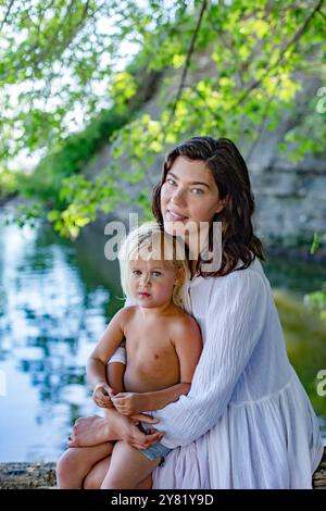 Une mère dans une robe blanche embrassant son jeune enfant par une rivière sereine avec un feuillage vert en arrière-plan. Banque D'Images